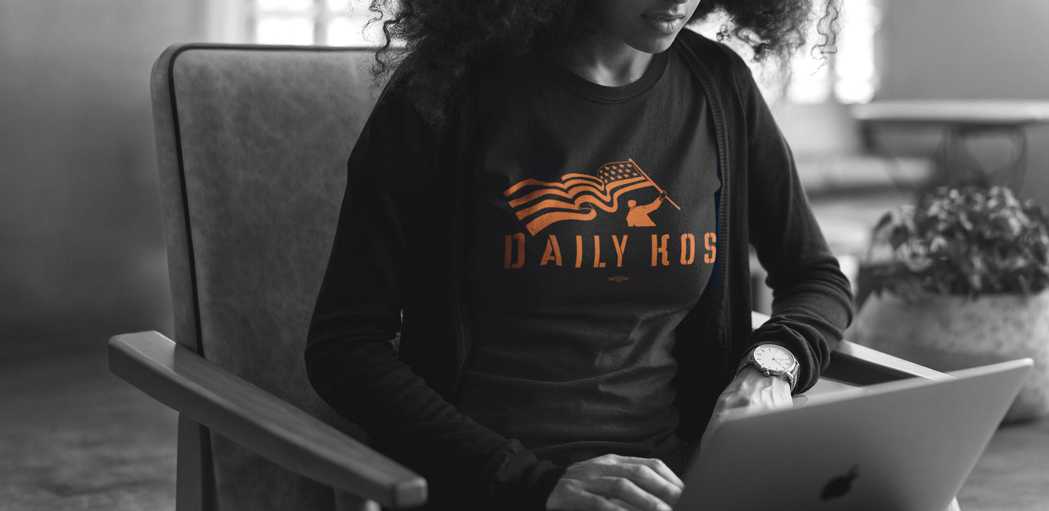 A person sitting in a chair using a laptop, wearing a black long-sleeve shirt with an orange graphic that reads 'DAILY KOS' and an American flag design. The photo is in black and white, with a blurred indoor background.